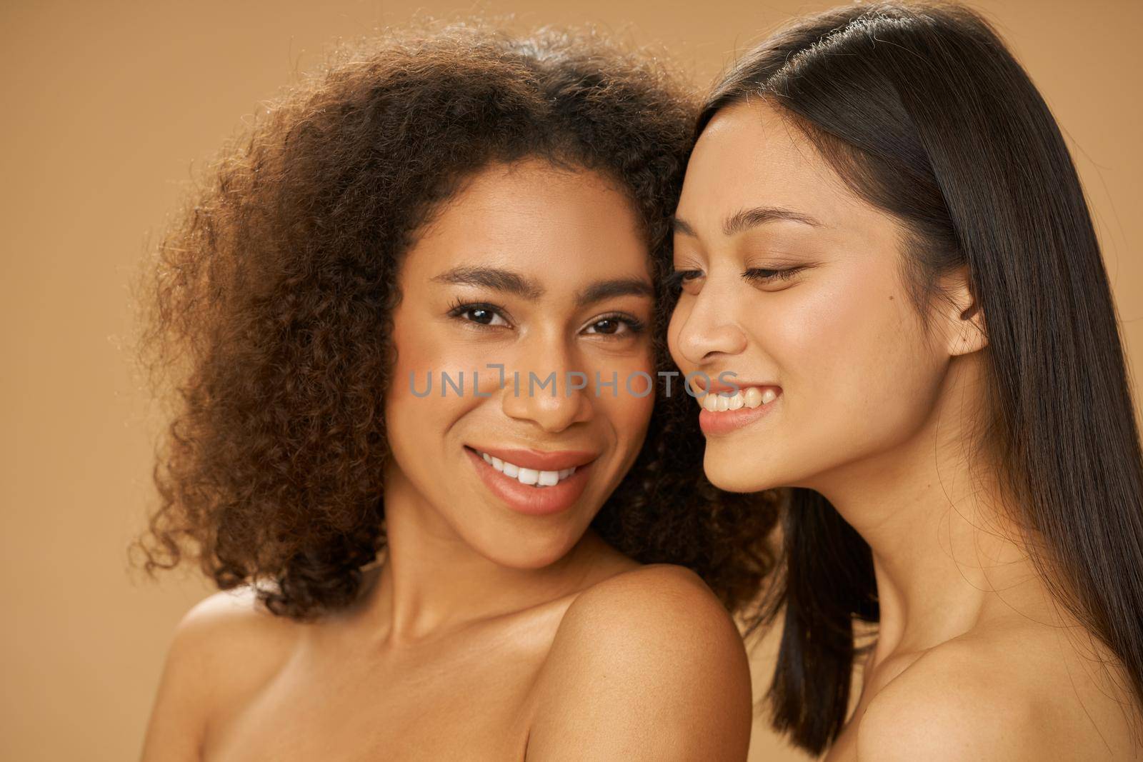 Portrait of two pretty mixed race young women with perfect smile posing together isolated over beige background. Health and beauty, diversity concept. Selective focus