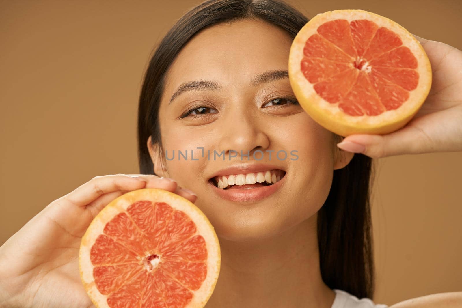 Beauty portrait of charming mixed race young woman smiling at camera, holding grapefruit cut in half, posing isolated over beige background. Health and beauty concept