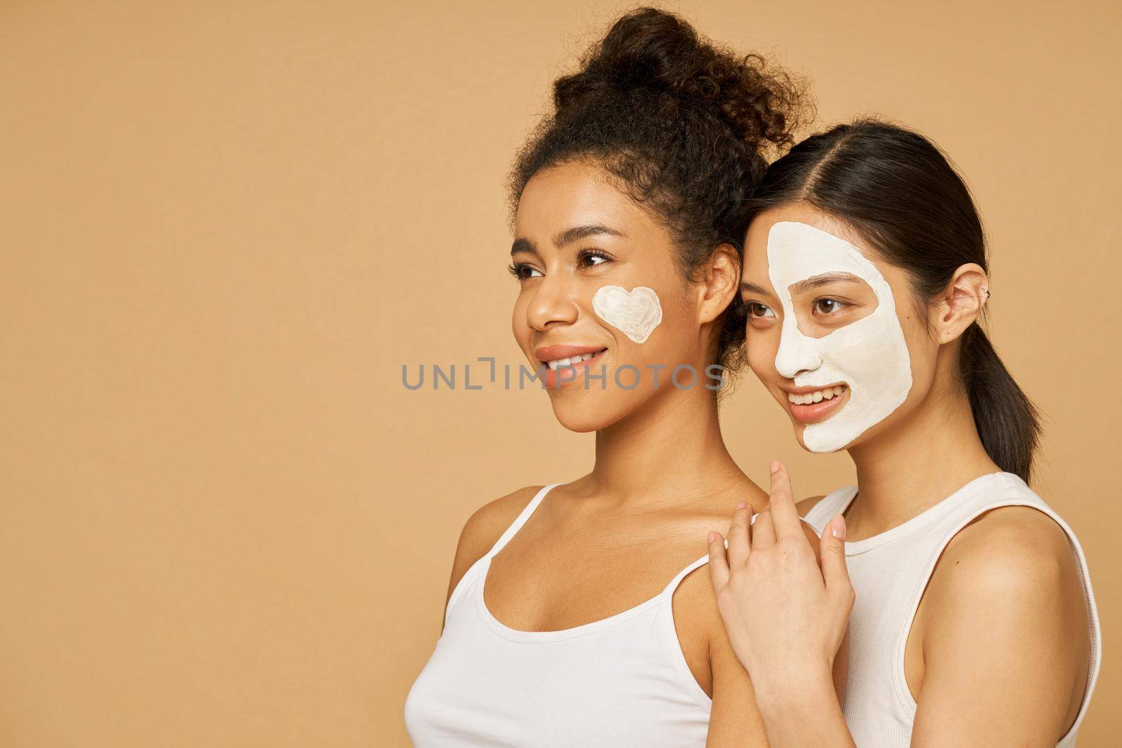 Portrait of two young women, female friends smiling aside while posing together with facial masks on isolated over beige background. Skincare, beauty concept