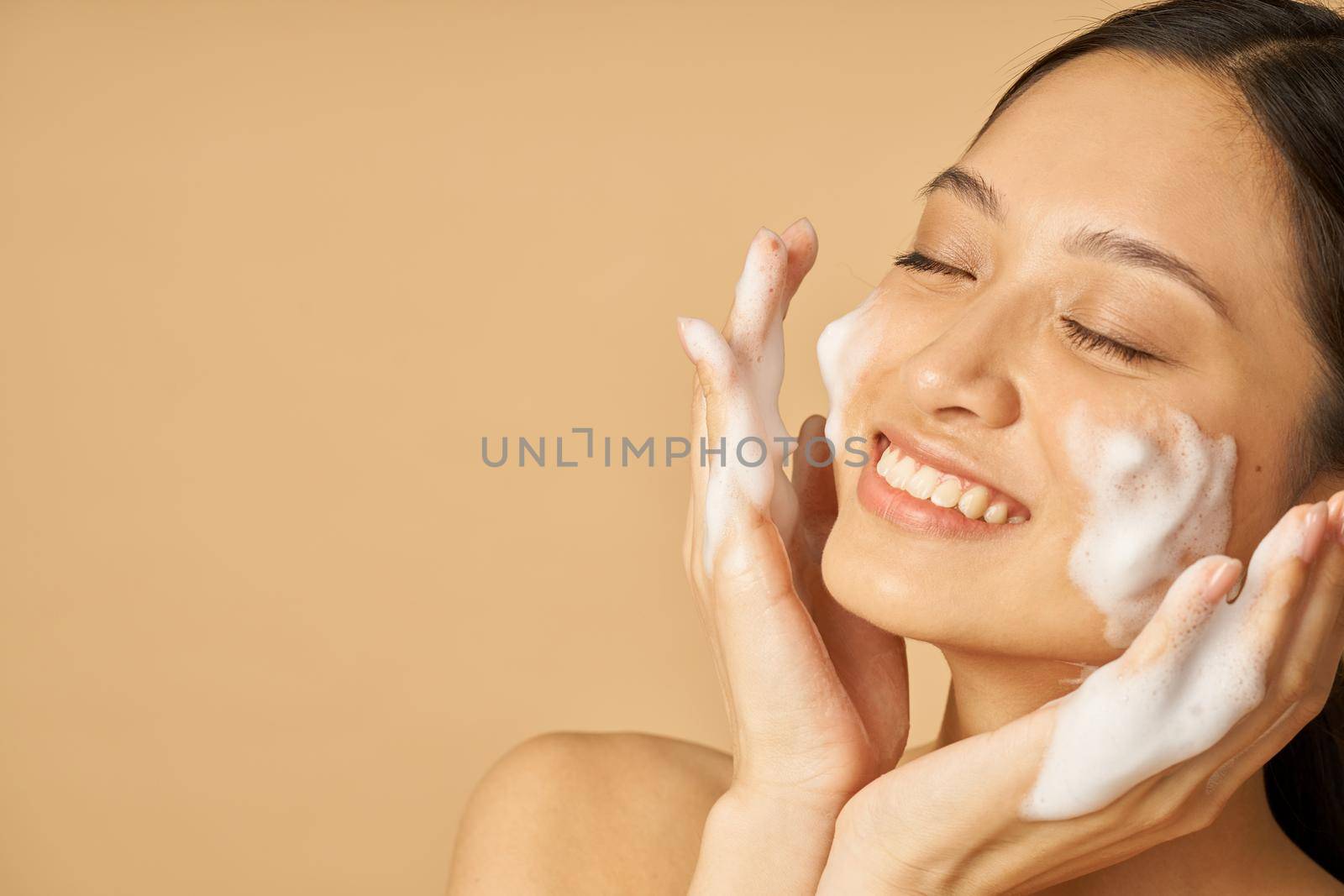 Beauty portrait of lovely young woman smiling with eyes closed while applying gentle foam facial cleanser isolated over beige background by friendsstock