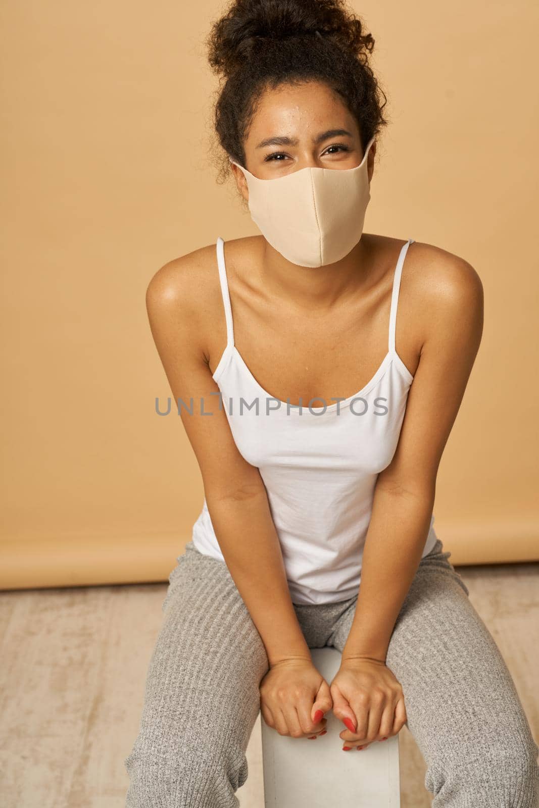 Studio shot of young mixed race woman in casual clothes wearing protective facial mask smiling at camera while posing over beige background by friendsstock