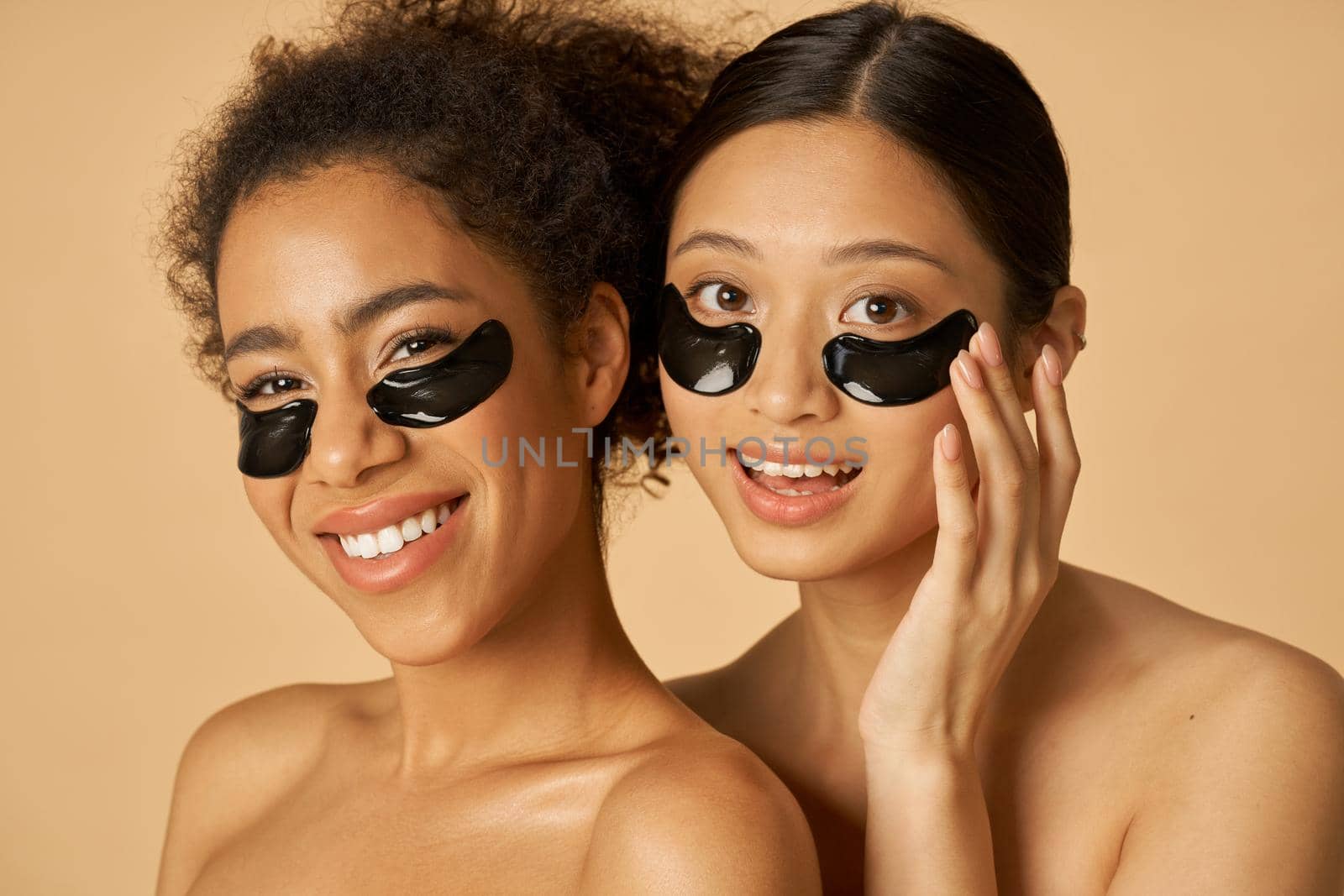 Beauty portrait of two happy young women posing with applied black under eye patches isolated over beige background by friendsstock