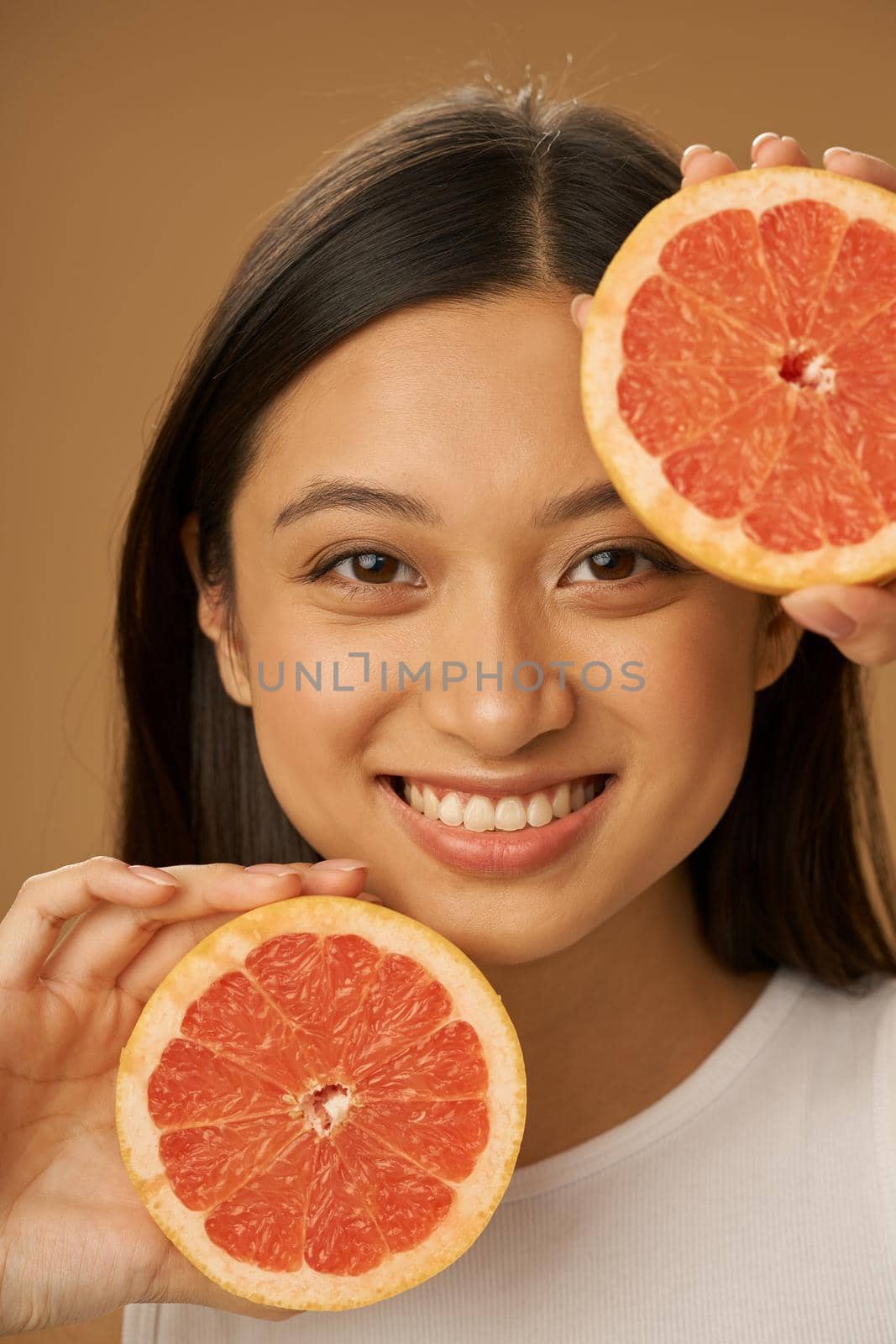 Portrait of excited mixed race young woman smiling at camera, holding grapefruit cut in half while posing isolated over beige background by friendsstock