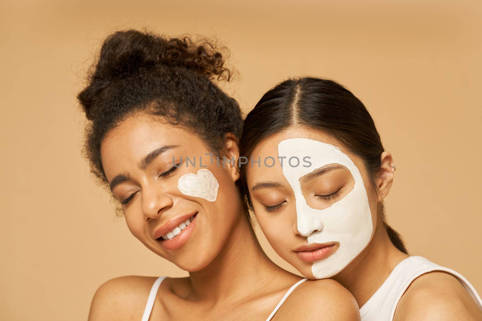 Close up portrait of two young female friends with facial masks on posing with eyes closed isolated over beige background. Skincare, beauty concept