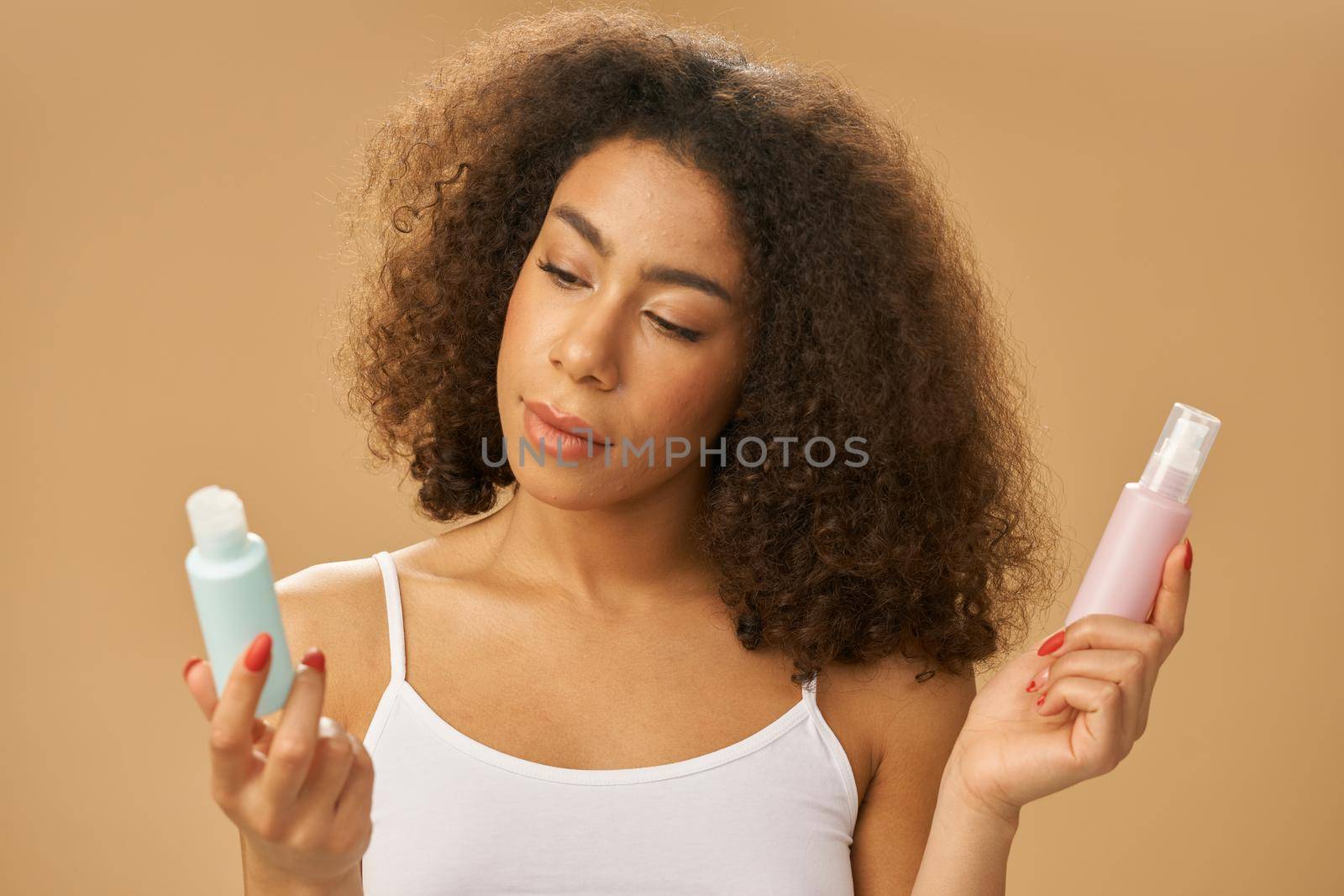 Portrait of attractive healthy african american young woman choosing between two bottles with beauty product while posing over beige background by friendsstock