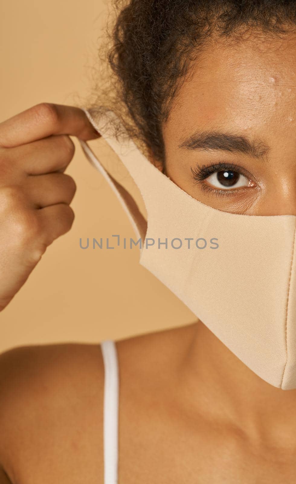 Half face portrait of young mixed race woman adjusting her facial mask, looking at camera, posing over beige background by friendsstock