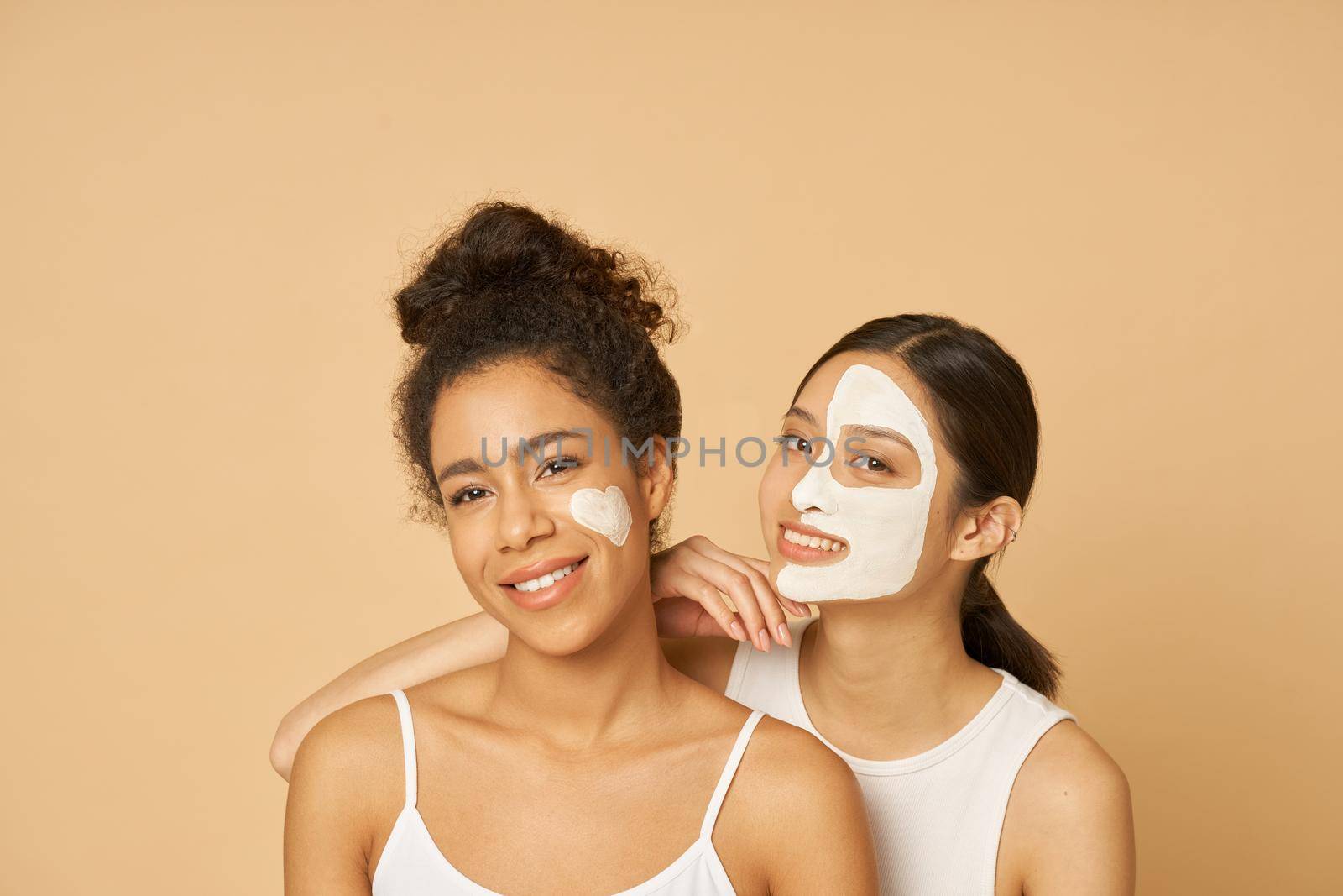 Studio shot of two young women, female friends having fun, smiling at camera while posing with facial masks on isolated over beige background by friendsstock