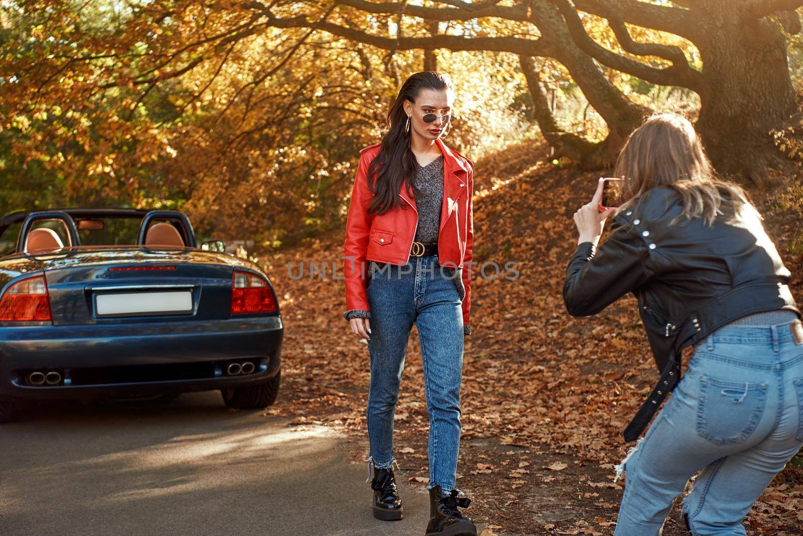 Woman photographing her friend in red coat and sunglasses near cabriolet by friendsstock