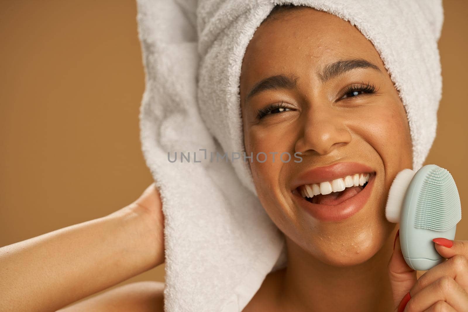 Face closeup of joyful young woman after shower looking excited at camera while using silicone facial cleansing brush, posing isolated over beige background. Beauty, spa treatment concept