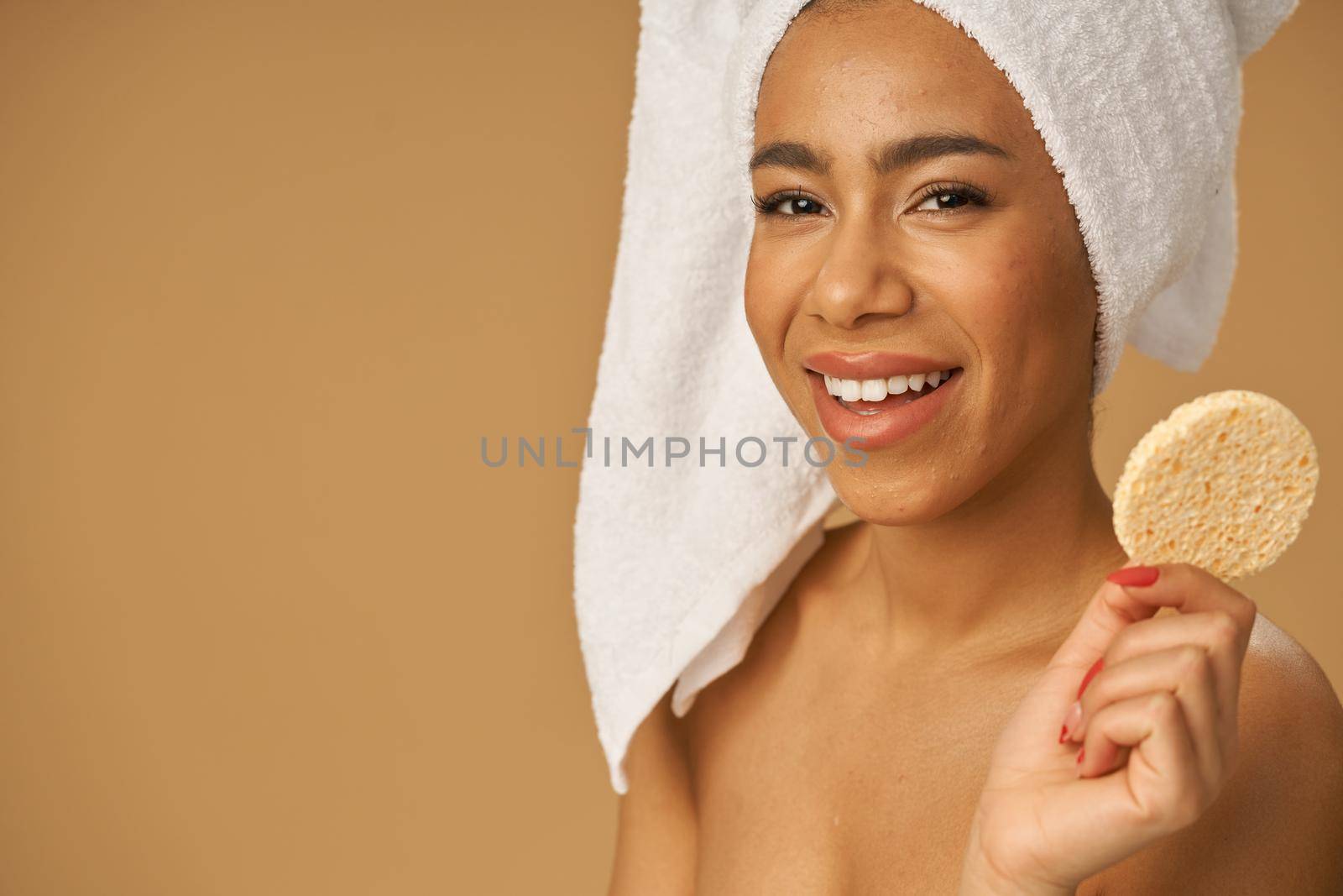 Joyful mixed race young woman smiling at camera, holding cleansing face sponge, posing isolated over beige background. Bathroom routine. Youth and skin care concept. Horizontal shot