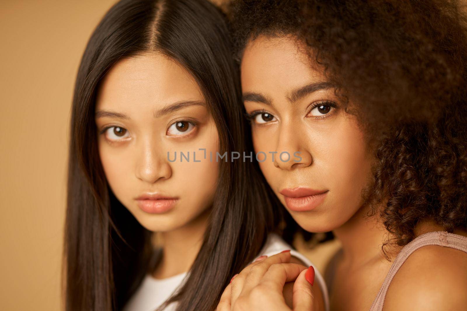 Face closeup of two beautiful mixed race young women with perfect skin looking at camera while posing isolated over beige background. Female beauty portrait