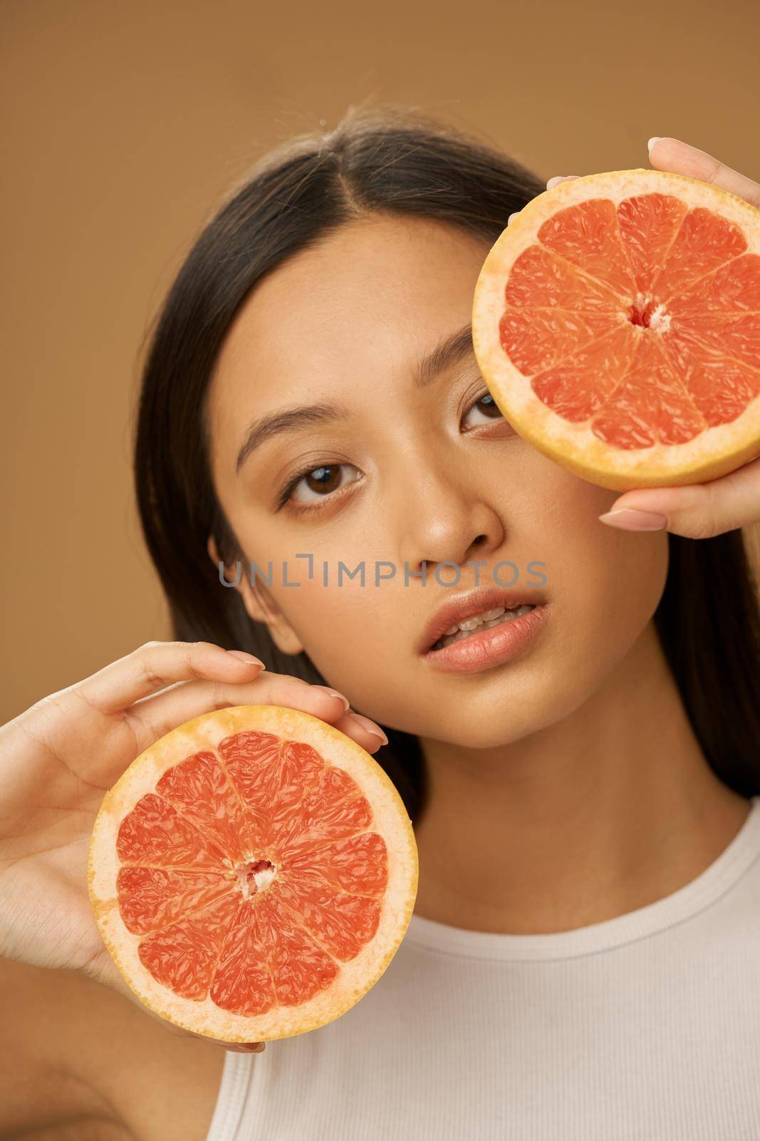 Beauty portrait of natural mixed race young woman holding grapefruit cut in half, posing isolated over beige background. Health and beauty concept