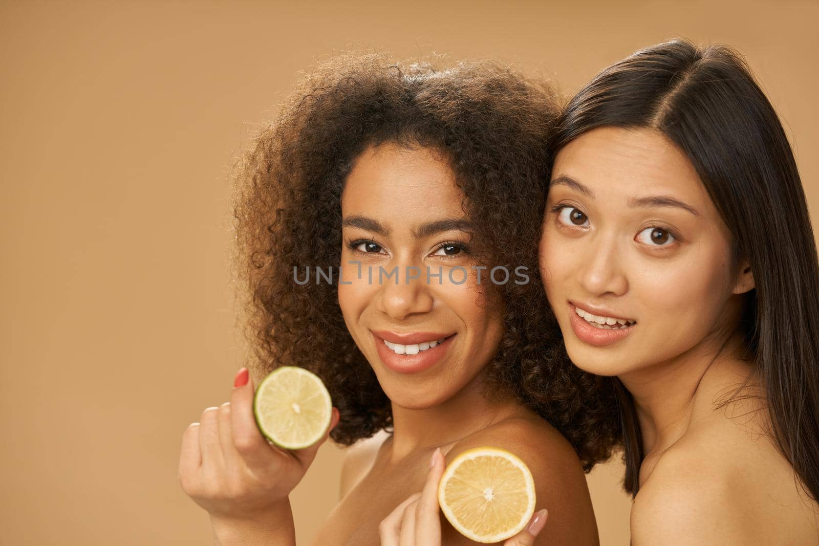 Two cute mixed race young women looking at camera and holding lemon and lime cut in half while posing together isolated over beige background by friendsstock