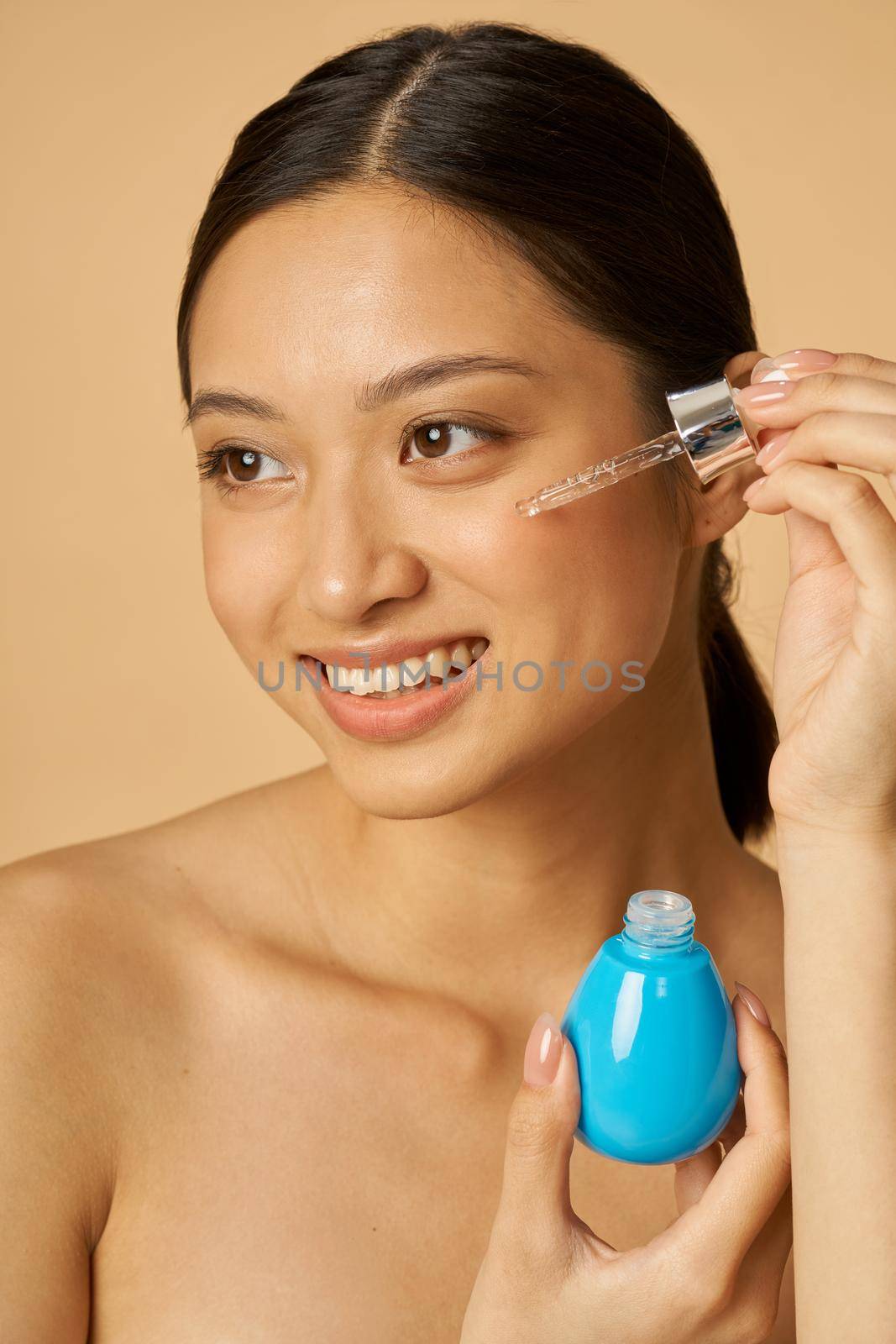 Portrait of pretty young woman dropping oil serum with pipette on face for fresh perfect skin, posing isolated over beige background by friendsstock