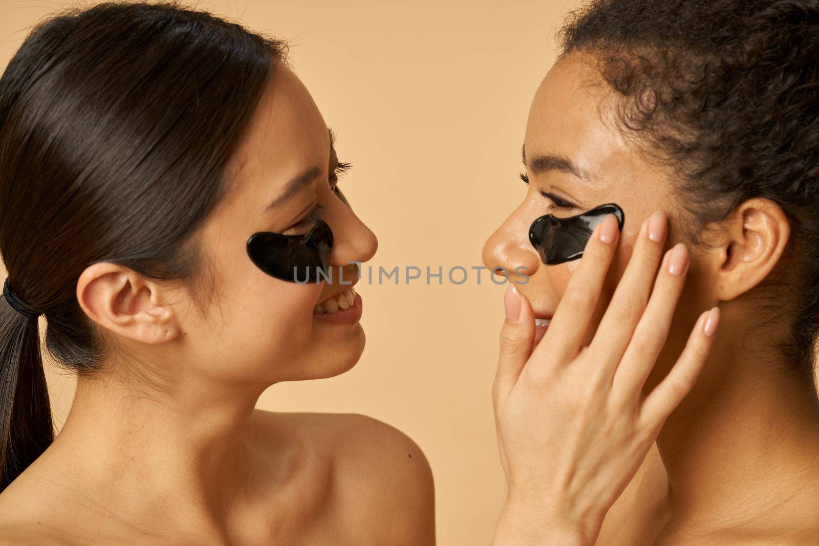 Close up portrait of adorable young women applying black hydro gel under eye patches on facial skin, posing isolated over beige background by friendsstock