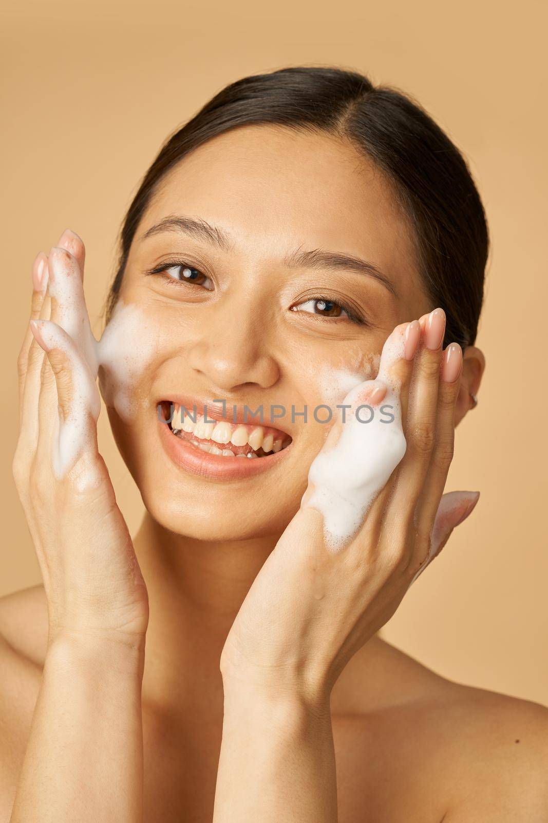 Beauty portrait of joyful young woman smiling at camera while applying gentle foam facial cleanser isolated over beige background by friendsstock