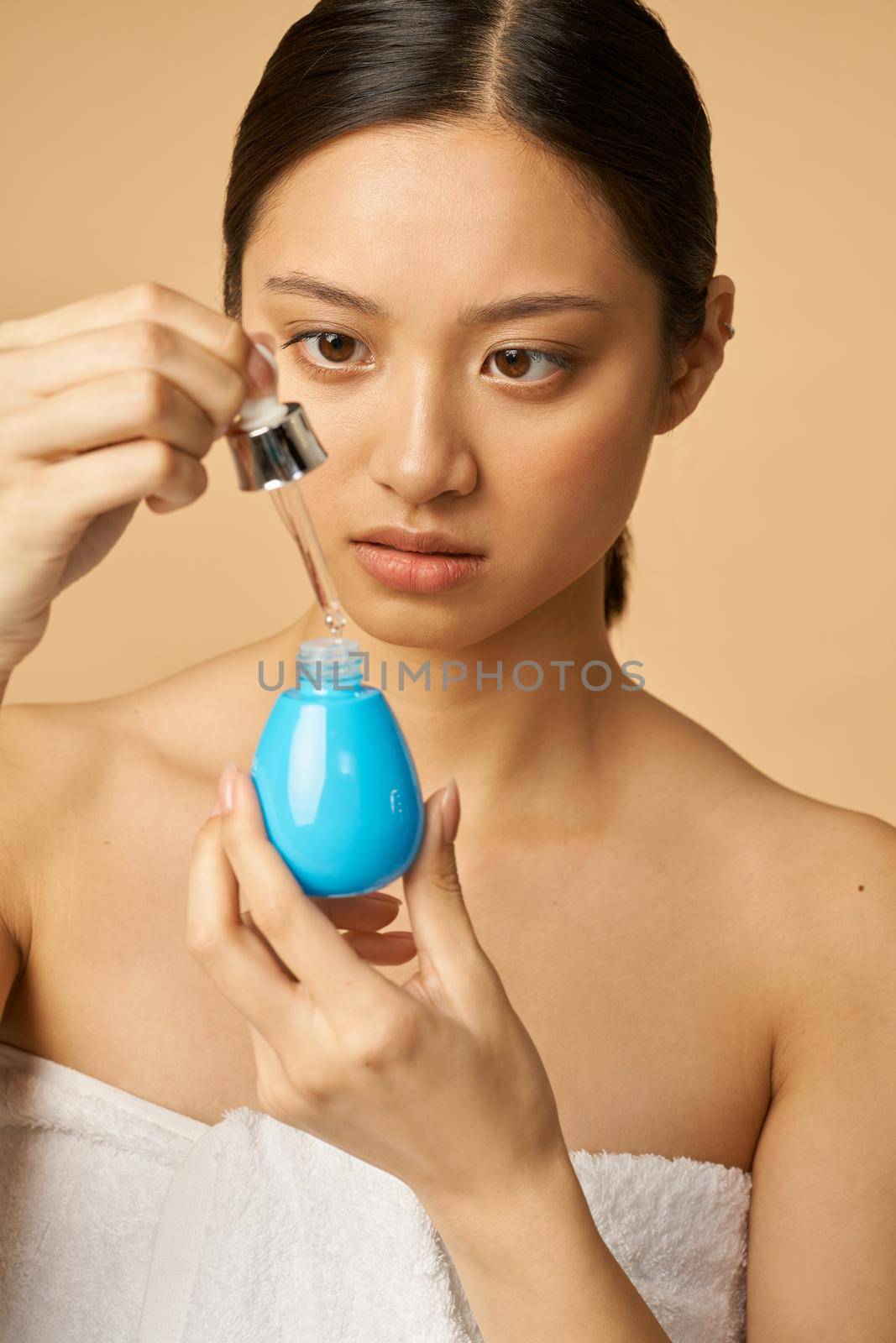 Portrait of young woman looking focused while holding oil drop serum, posing isolated over beige background. Beauty treatment concept