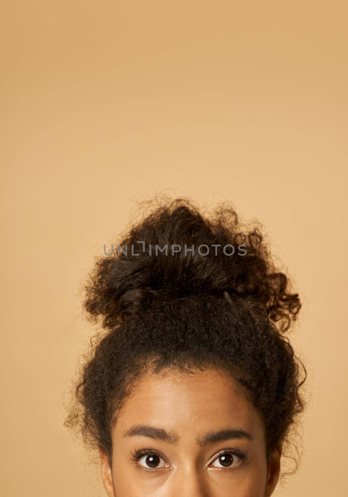 Cropped portrait of young mixed race woman with highly raised dark curly hair looking at camera while posing isolated over beige background. Vertical shot