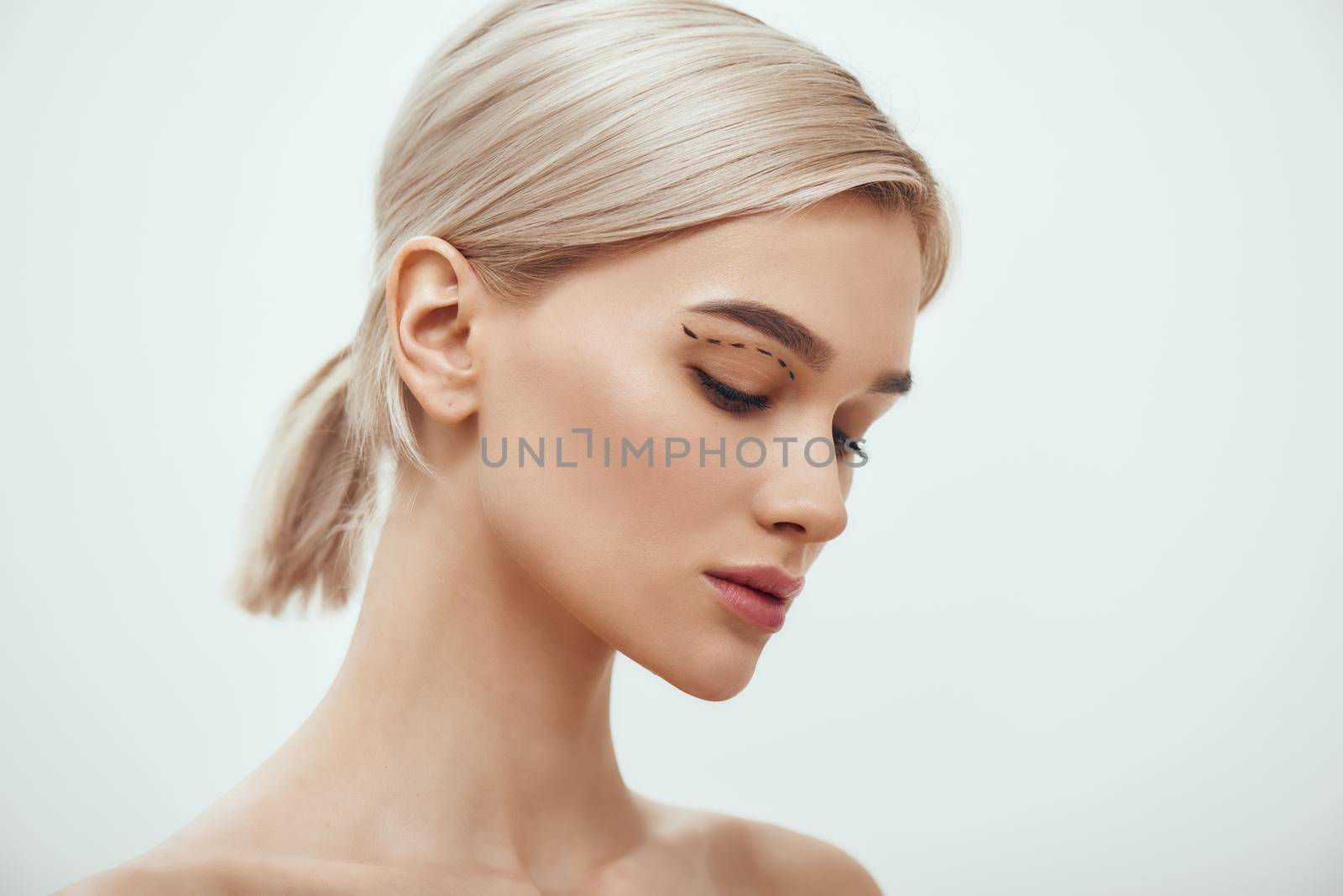 Before facial surgery. Side view of pretty young blonde woman with sketch on her face standing against grey background by friendsstock