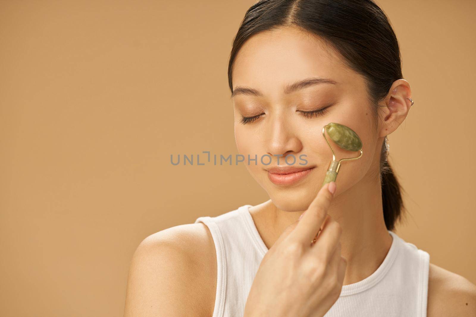 Facial beauty treatment. Beautiful young woman looking relaxed while getting massage face using jade roller isolated over beige background by friendsstock