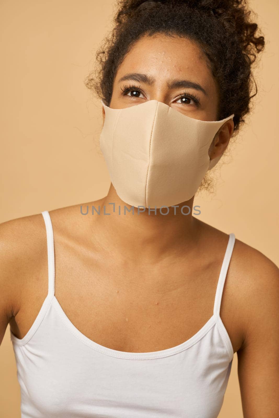 Portrait of adorable young mixed race woman wearing protective facial mask looking aside while posing isolated over beige background by friendsstock