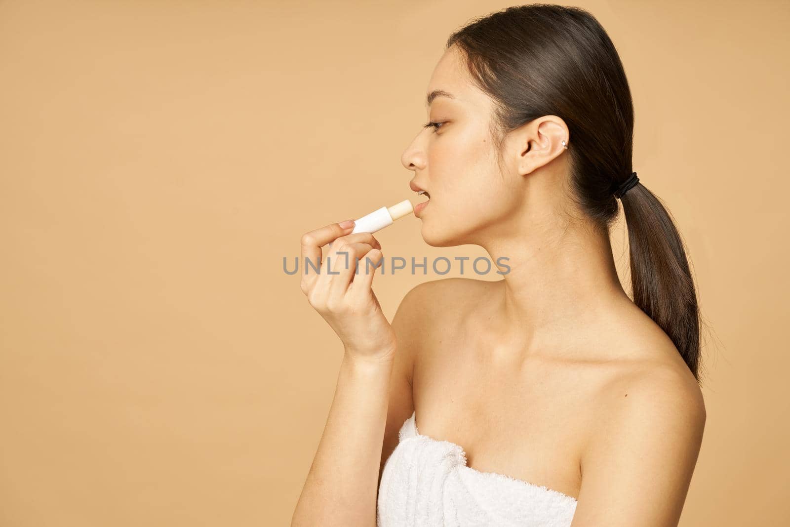 Charming young woman wrapped in towel looking aside while applying lip balm, posing isolated over beige background by friendsstock