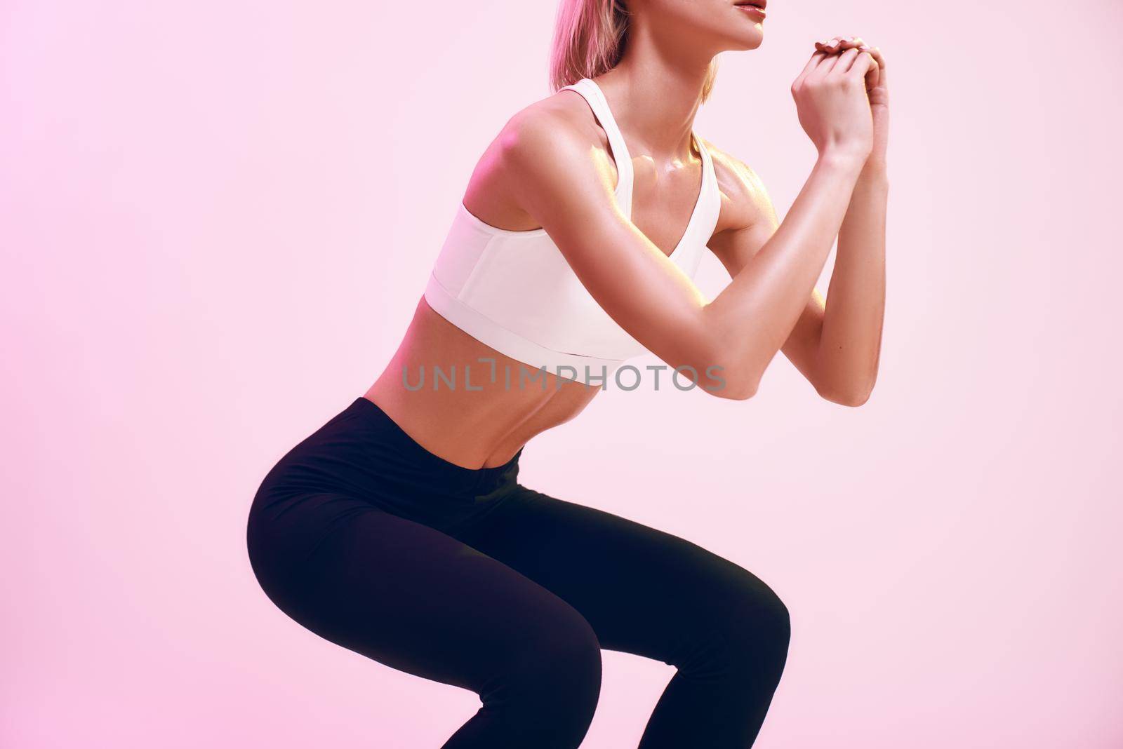 Perfect squat. Cropped photo of sporty and cute slim woman in sportswear doing squat while standing against pink background in studio. Sport. Active lifestyle. Workout