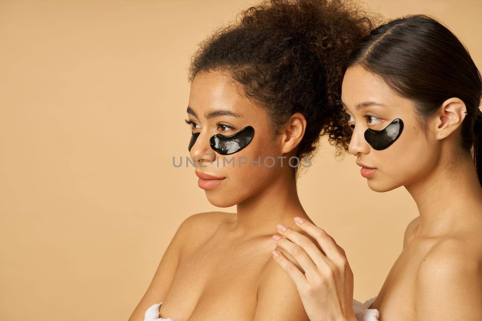 Beauty portrait of two happy young women looking away, posing with applied black hydro gel under eye patches isolated over beige background. Skincare routine concept