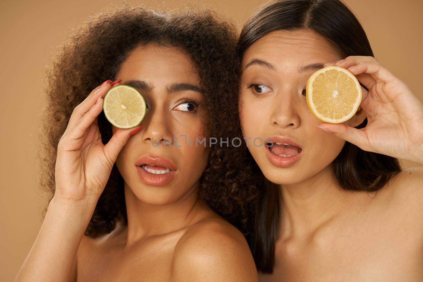 Portrait of two funny mixed race young women looking surprised, holding lemon and lime cut in half while posing together isolated over beige background. Health and beauty concept