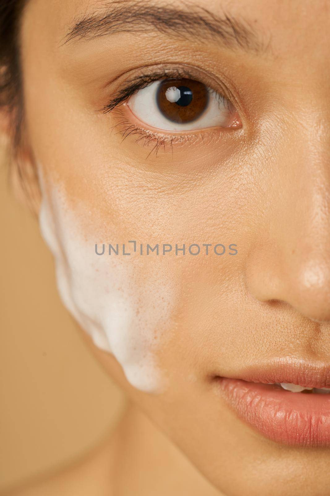 Close up half face portrait of beautiful young woman looking at camera with gentle foam facial cleanser applied on her face isolated over beige background. Beauty products and skin care concept