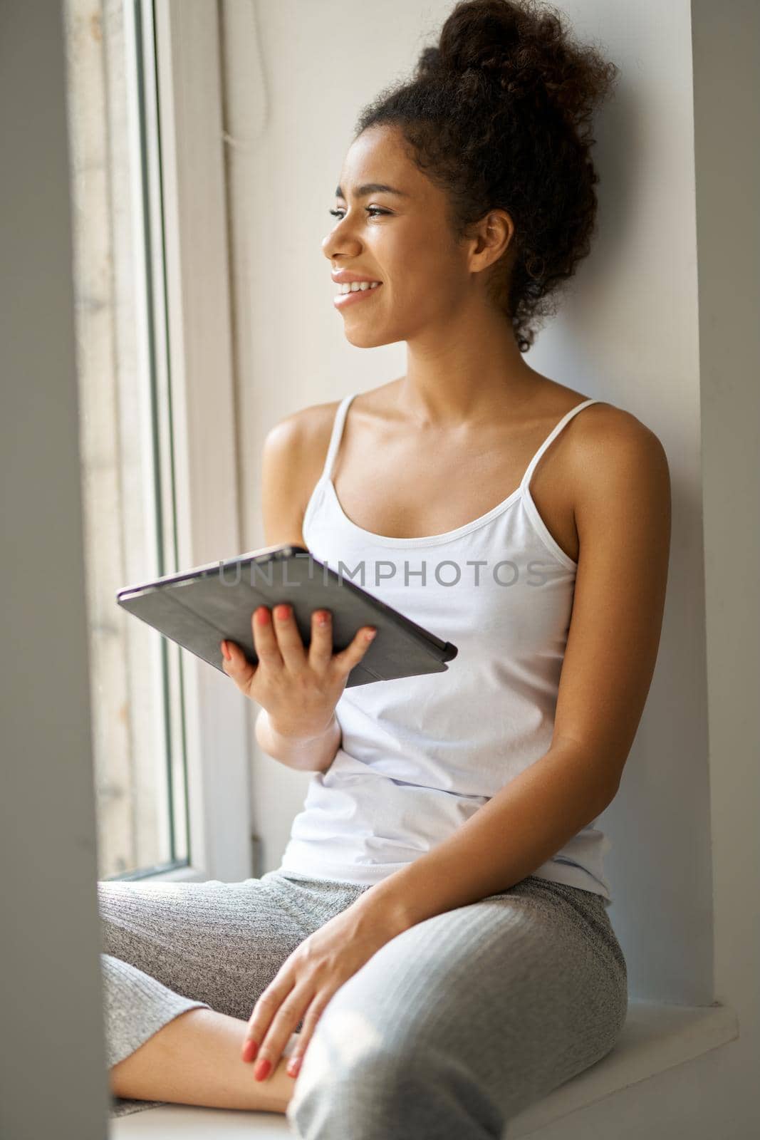 Young mixed race woman with tablet pc looking out the window, feeling cozy while sitting alone by the window at home. Lifestyle, weekend concept