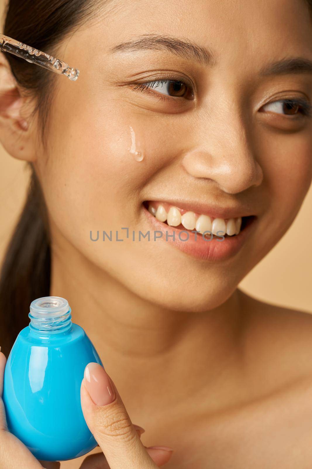 Portrait of pretty young woman smiling aside, dropping oil serum with pipette on face for fresh perfect skin isolated over beige background by friendsstock