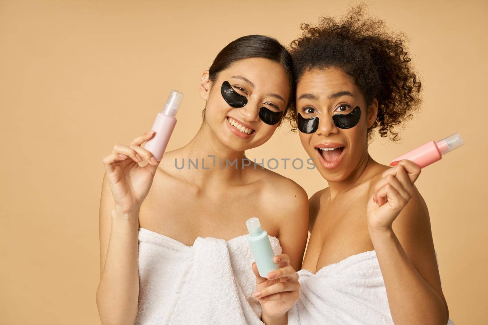 Two cute, young women with hydro gel under eye patches looking excited, holding bottles with beauty product, posing isolated over beige background by friendsstock