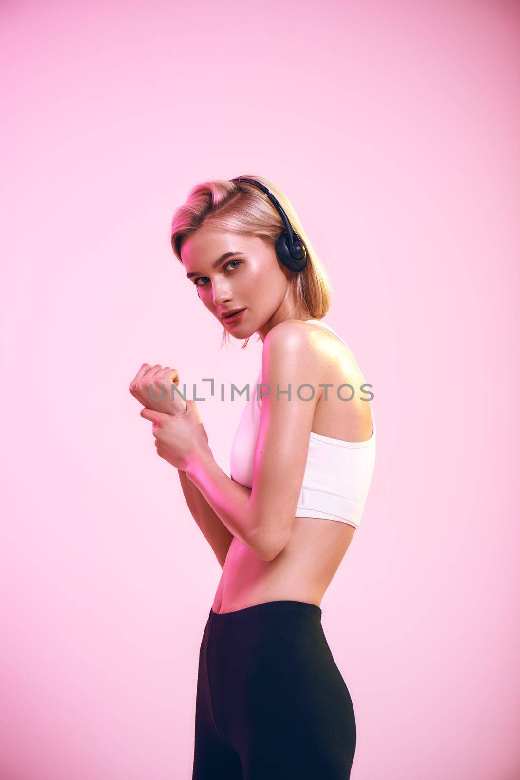 Music is my motivation. Side view of sporty slim blonde woman in headphones listening music while standing against pink background by friendsstock