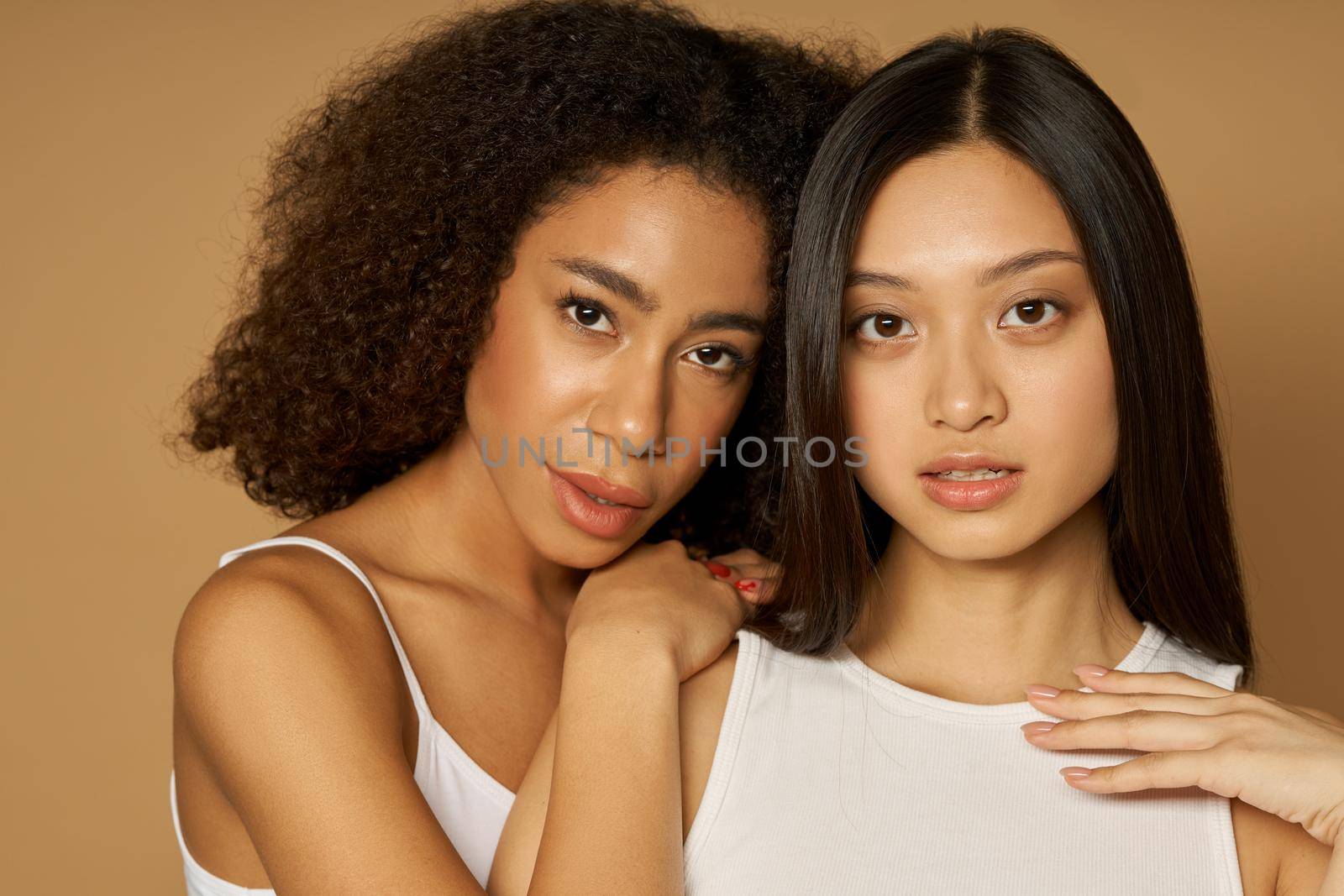 Beauty portrait of two mixed race young women with perfect skin looking at camera while posing together, standing isolated over light brown background. Skincare, diversity concept