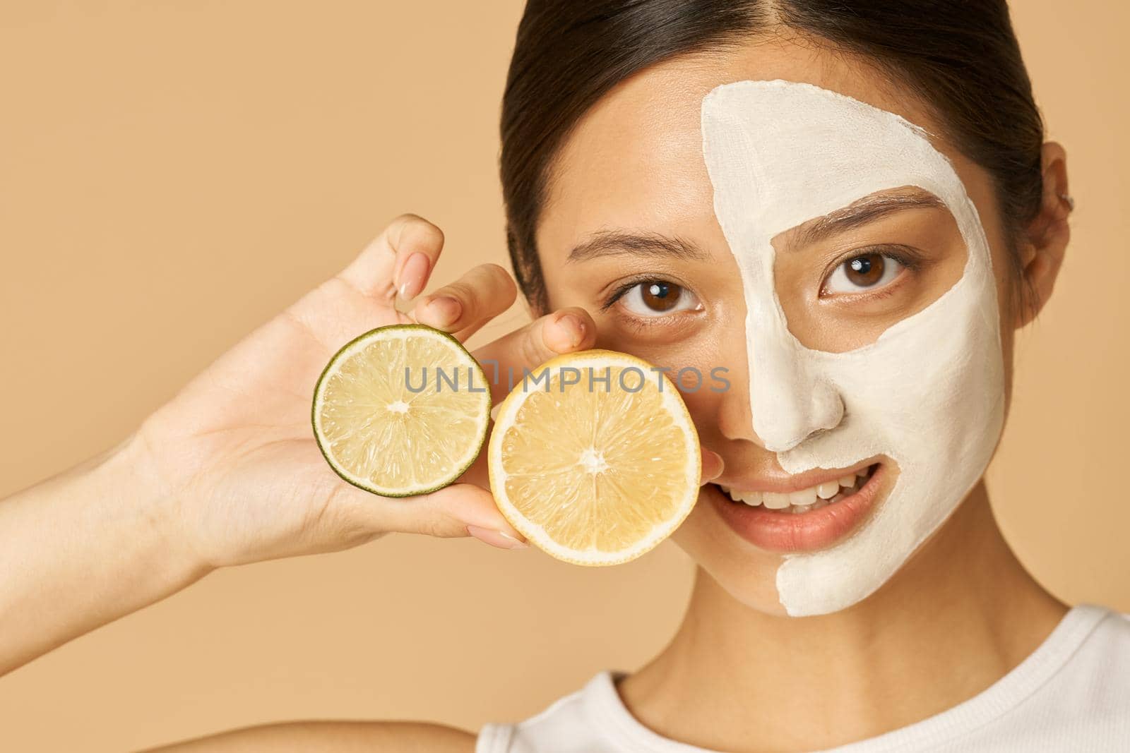 Young woman with facial mask applied on half of her face holding slice of lime and lemon, posing isolated over beige background by friendsstock