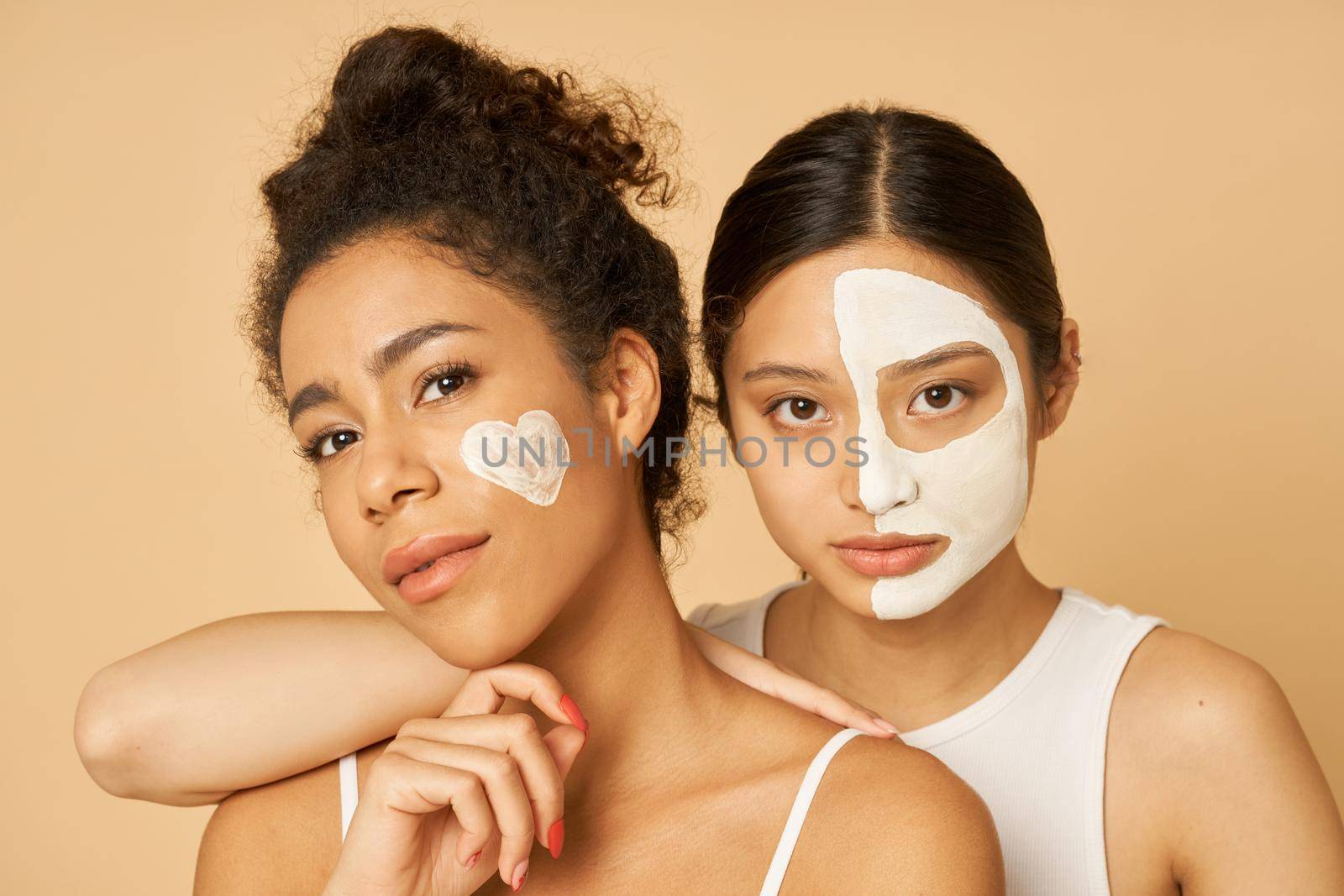 Portrait of two young women, female friends looking at camera while posing together with facial masks on isolated over beige background by friendsstock