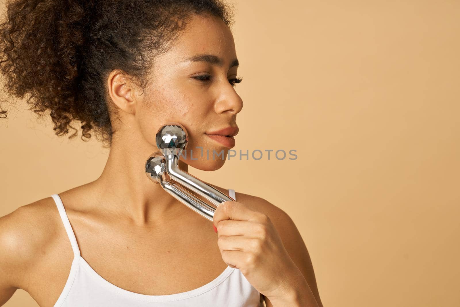 Portrait of attractive young woman looking aside while using silver metal face roller, posing isolated over beige background. Facial massage concept