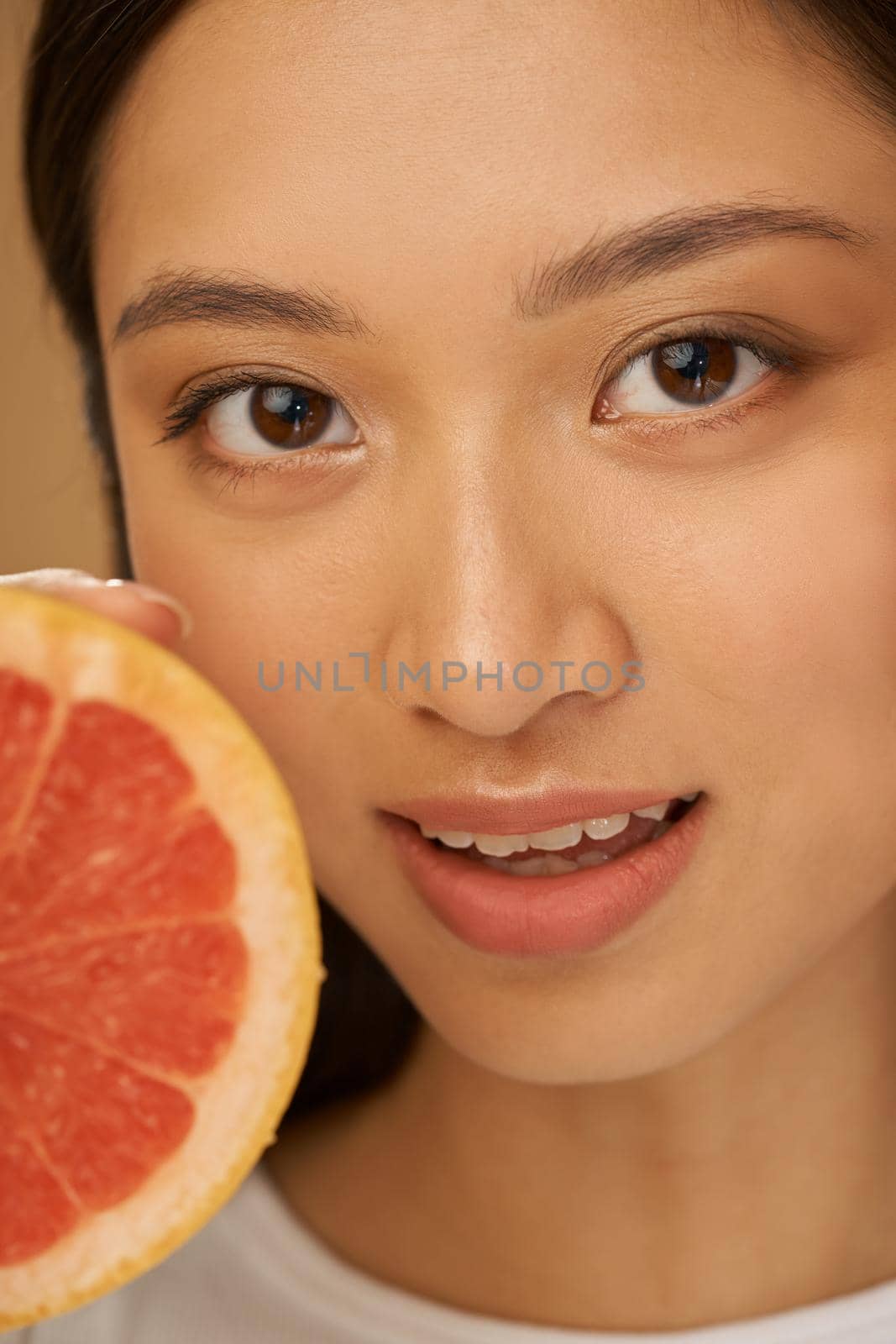 Close up portrait of attractive mixed race young woman looking at camera, holding grapefruit cut in half, posing isolated over beige background by friendsstock