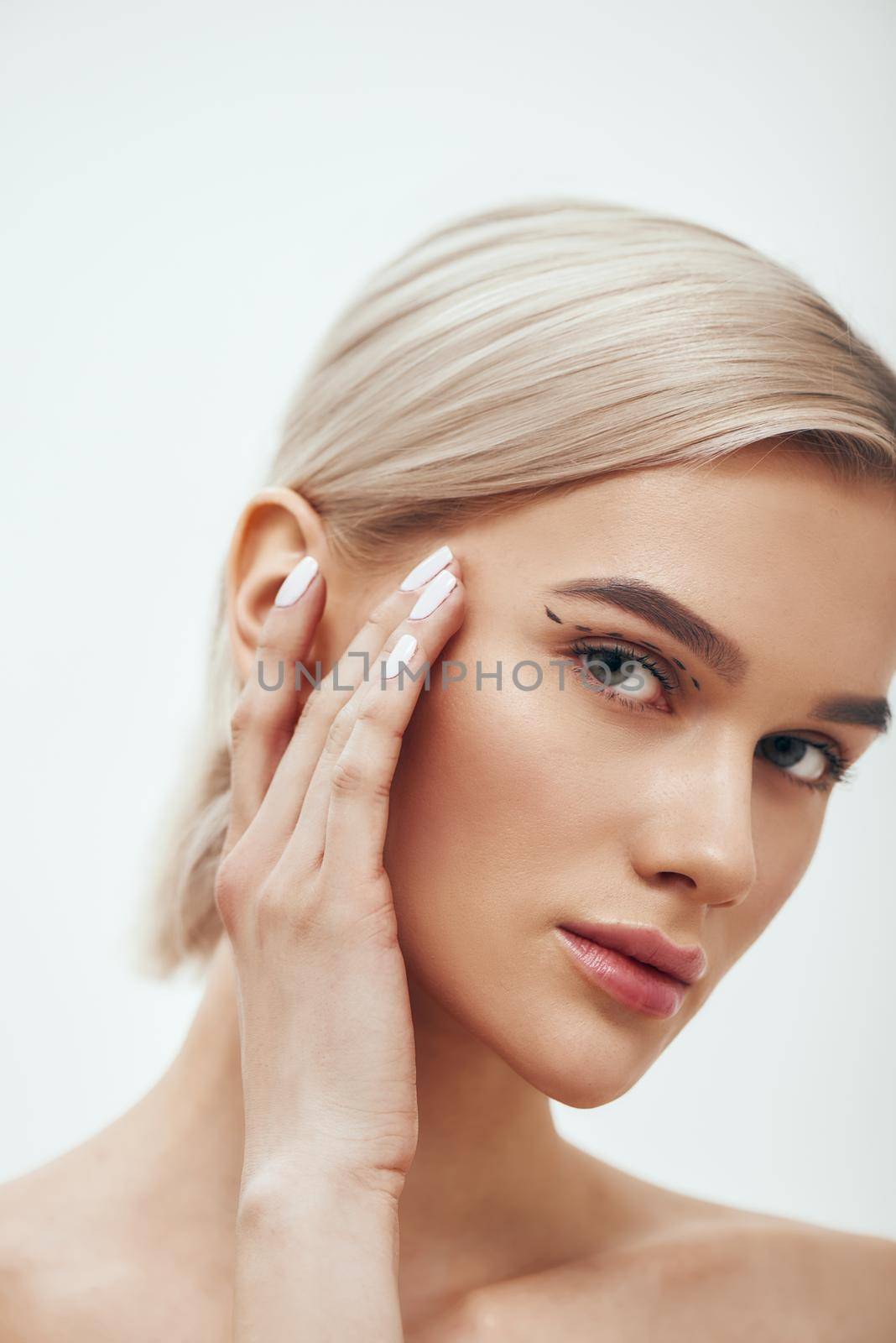 Pretty and young blonde woman touching her face with black surgical lines on eyelids and looking at camera. Plastic surgery concept by friendsstock