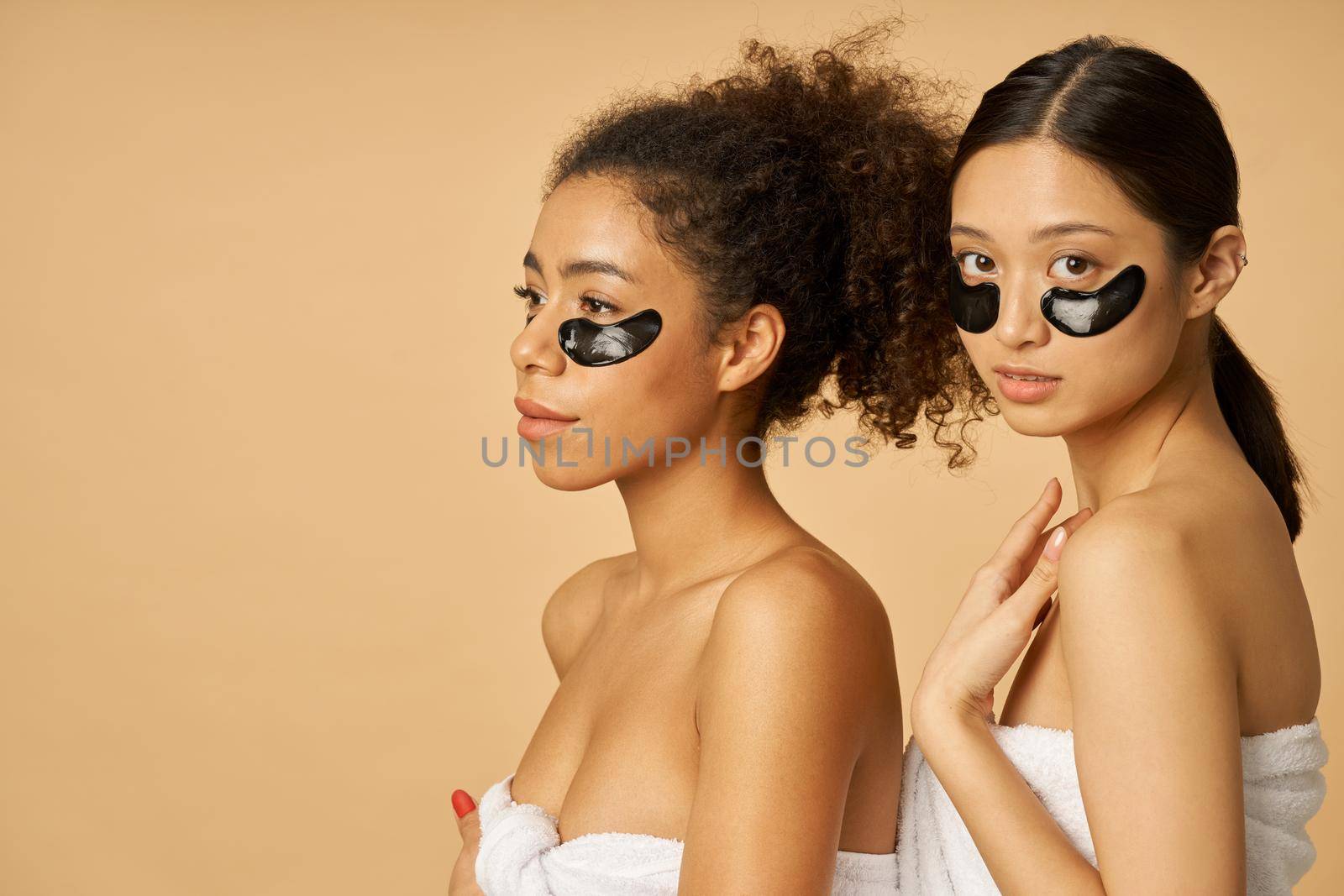 Two young women wrapped in towel posing with applied black hydro gel under eye patches isolated over beige background. Skincare routine concept