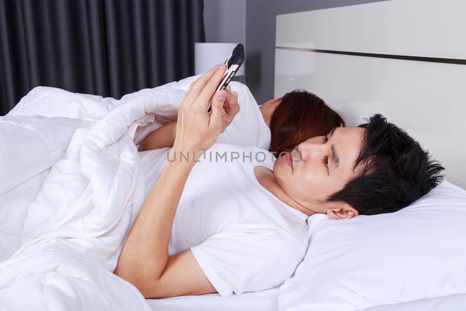 young man using his mobile phone in bed while his wife is sleeping next to him
