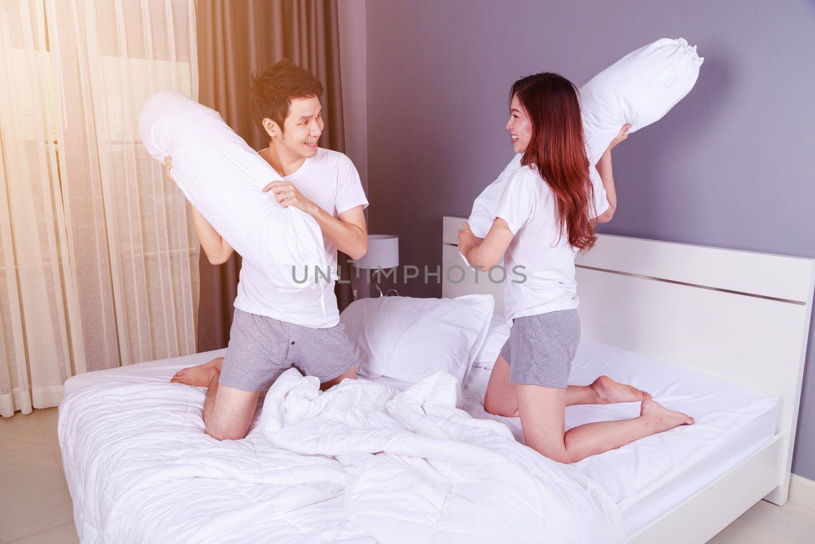 cheerful young couple having a bolster pillow fight on bed in bedroom by geargodz