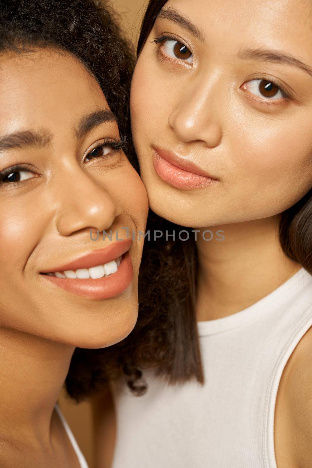 Face closeup of two beautiful mixed race young women with perfect glowing skin smiling at camera while posing together. Skincare, diversity concept. Selective focus. Vertical shot