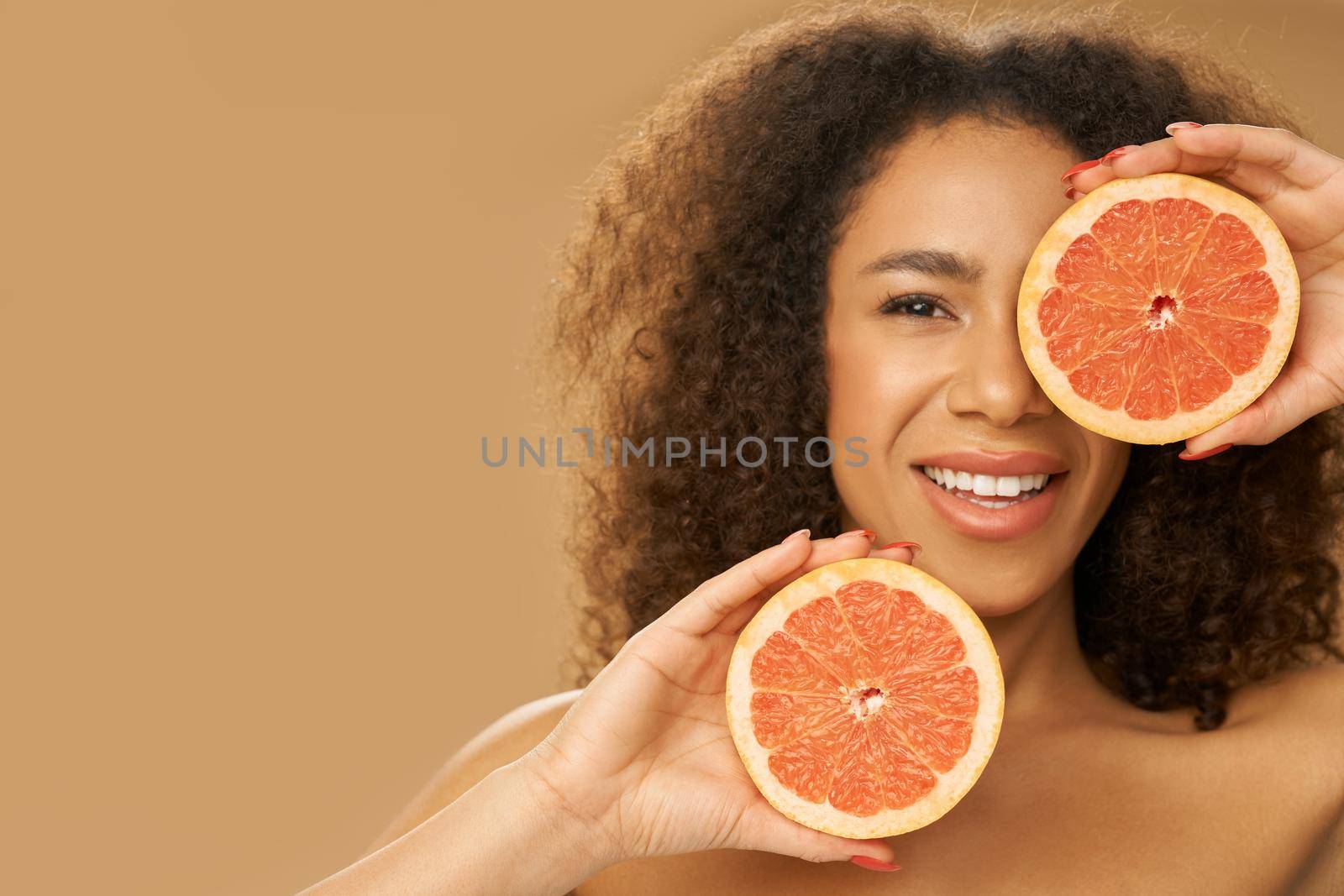 Portrait of joyful mixed race young woman covering one eye with grapefruit cut in half while posing isolated over beige background. Health and beauty concept. Front view