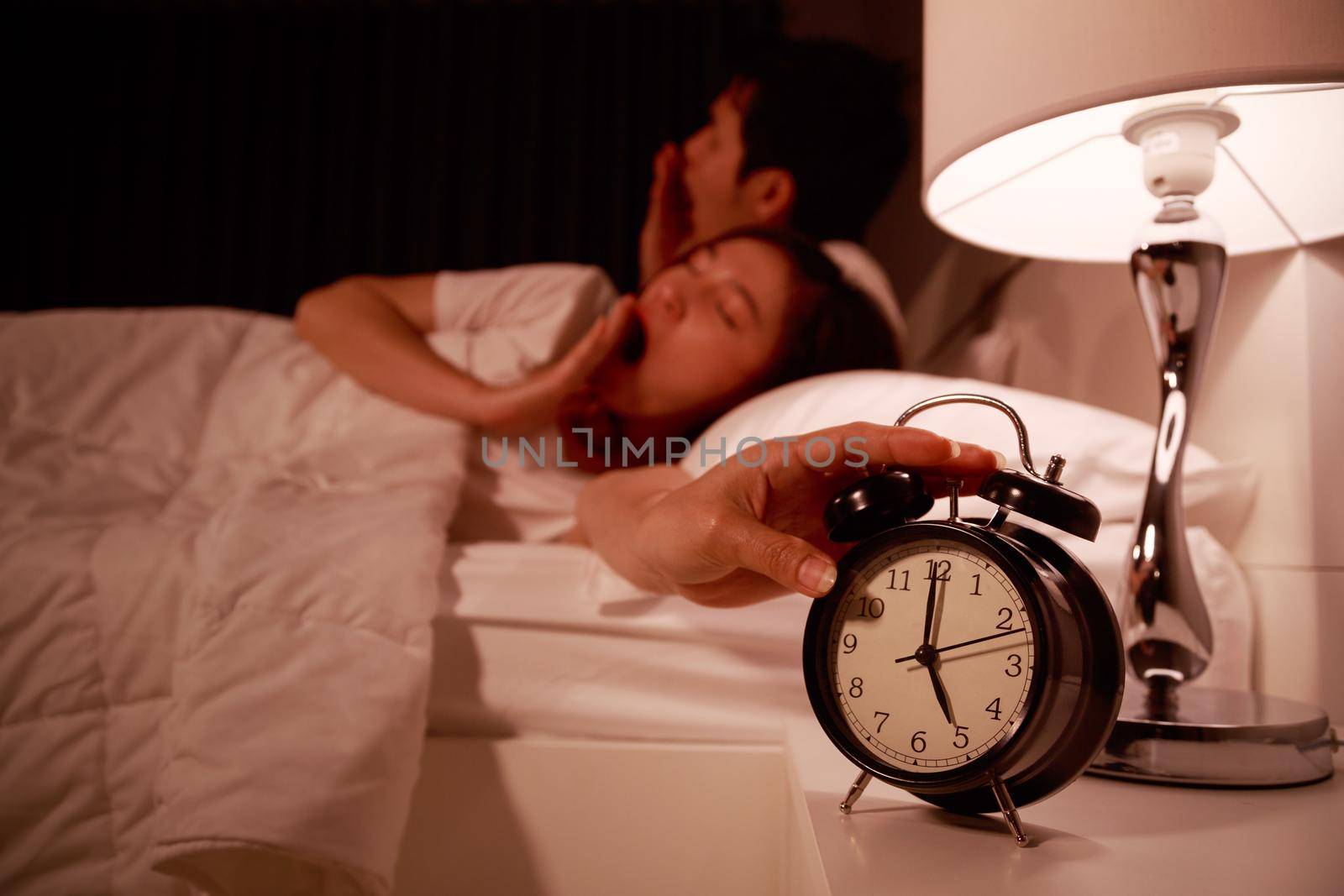 sleepy couple in bed with extending hand to alarm clock in the morning by geargodz
