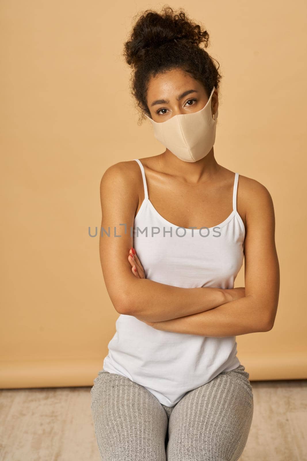 Studio shot of young mixed race woman in casual clothes wearing protective facial mask looking at camera while posing with arms crossed over beige background. Safety, pandemic concept