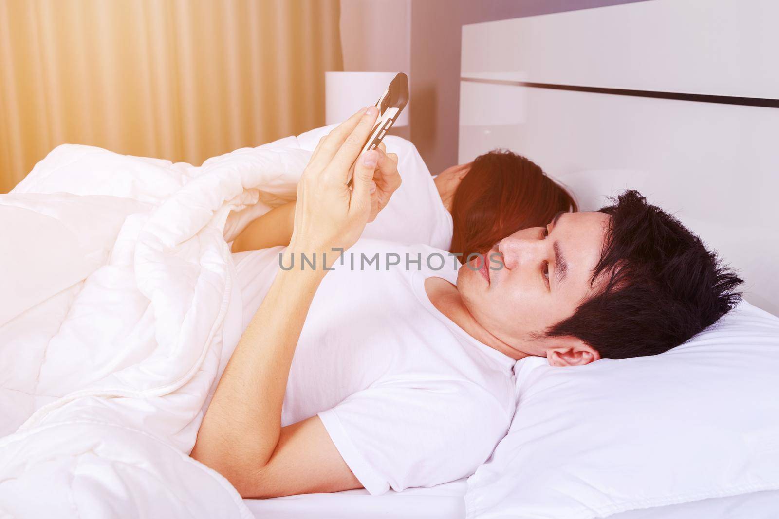 young man using his mobile phone in bed while his wife is sleeping next to him
