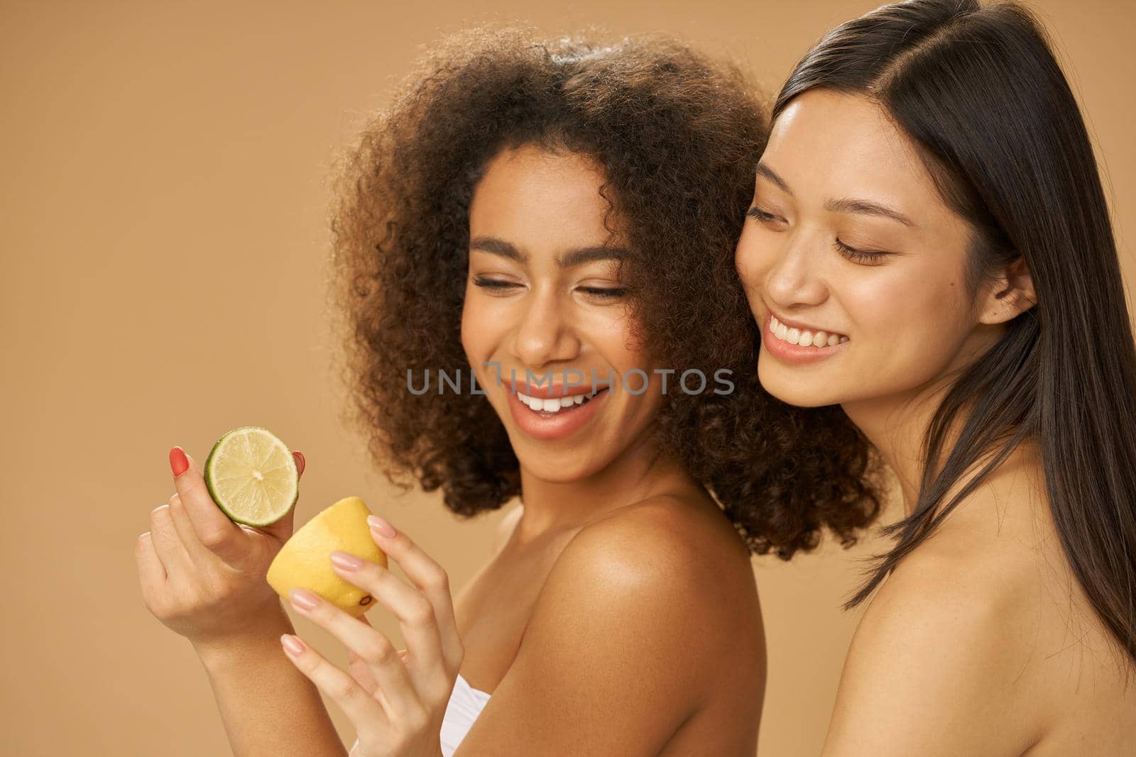 Two lovely mixed race young women looking happy, holding lemon and lime cut in half while posing together isolated over beige background by friendsstock