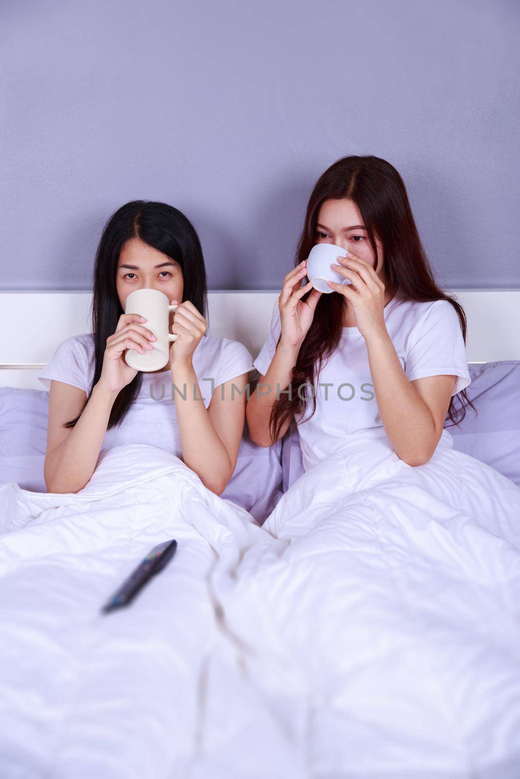 two best friends watching tv with remote and drinking a cup of coffee on bed in the bedroom 